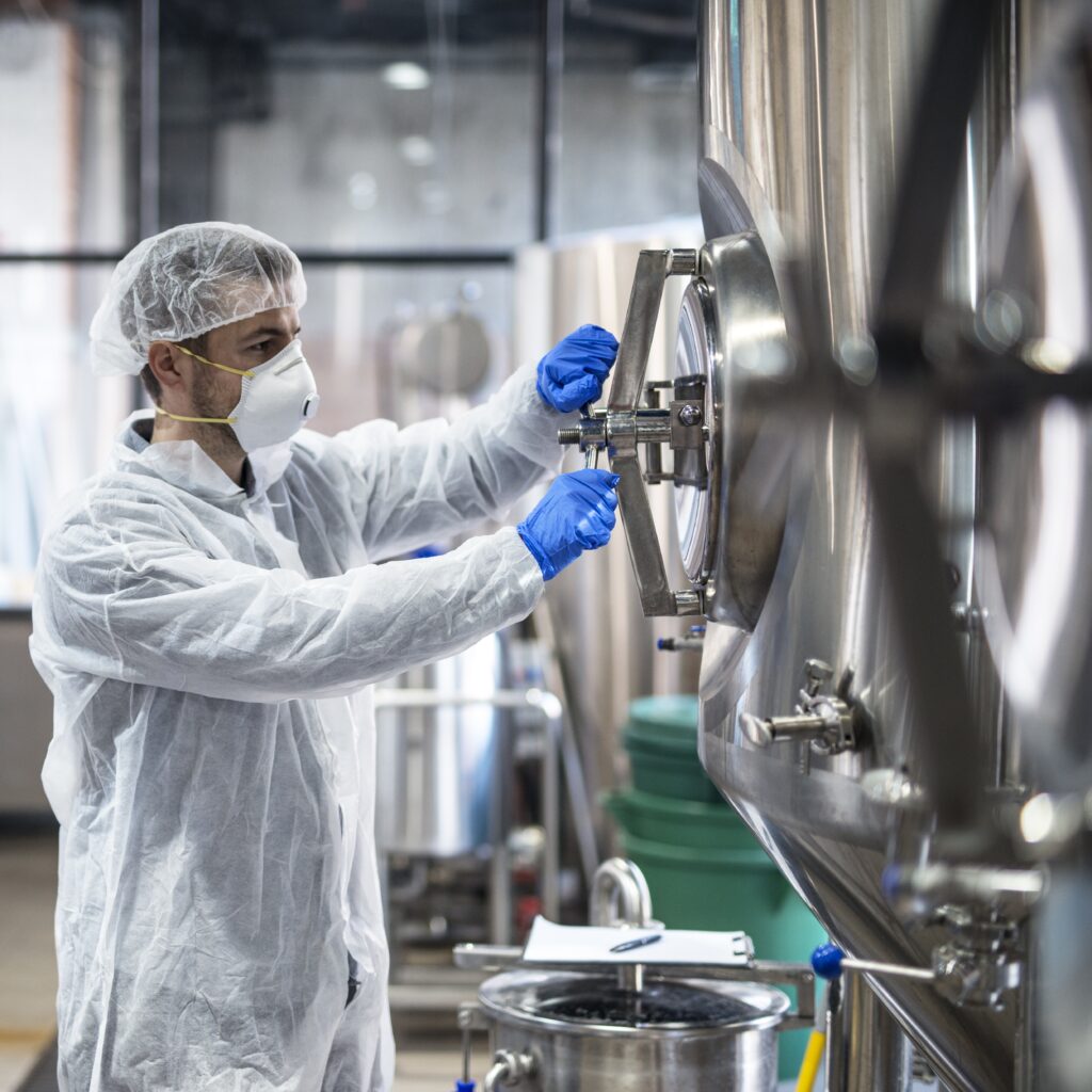 Technologist in protective uniform wearing hairnet mask goggles and gloves closing industrial tank reservoir.